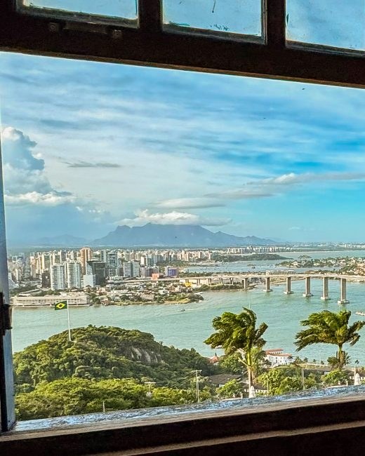 An old wooden window open onto the bay, with the Terceira Ponte bridge, the buildings of Vitória and the mountains beyond.
